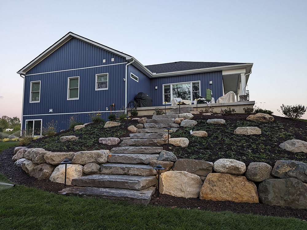 After: Image of the same home with stone slab steps, a boulder retaining wall, sod, and other greenery.